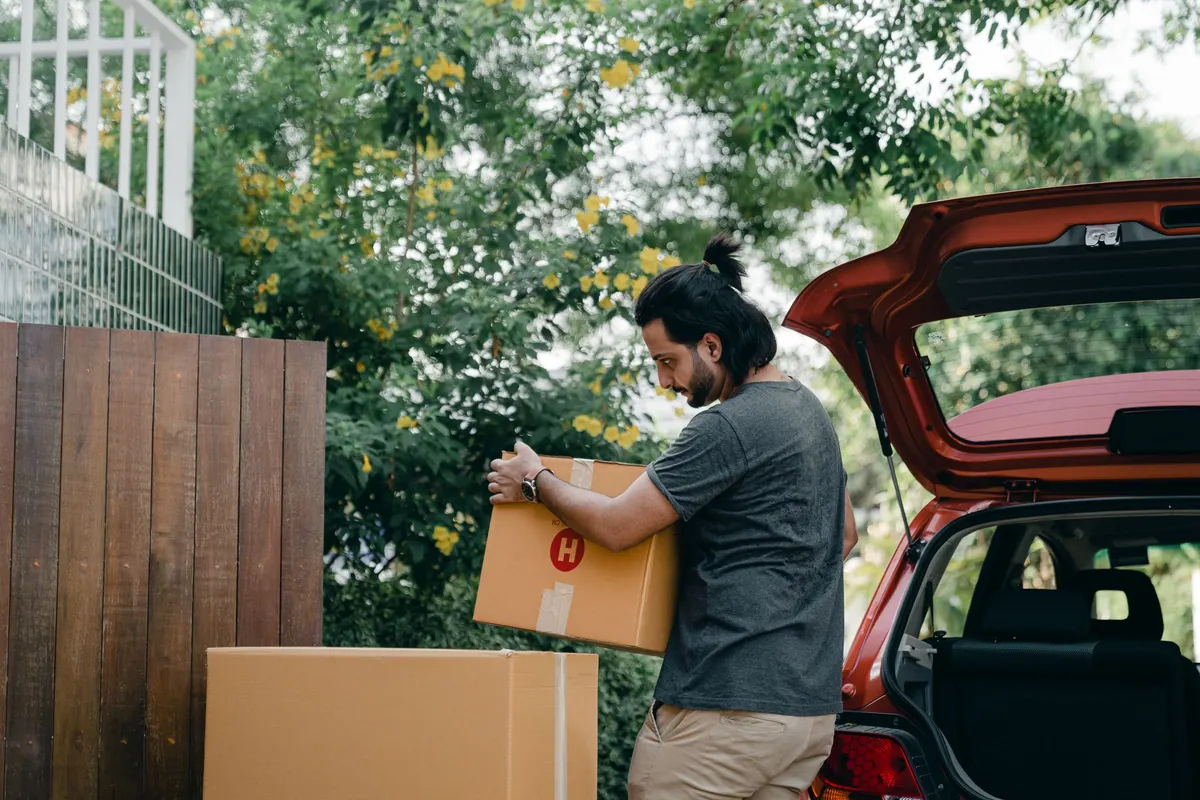 A man lifting a large taped cardboard box into the open boot of a car parked beside a garden wall.