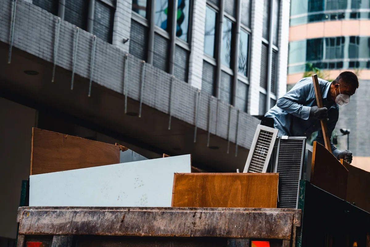 A worker in a face mask standing on an open-bed truck, loading large timber boards and metal grilles, with city office towers behind him.