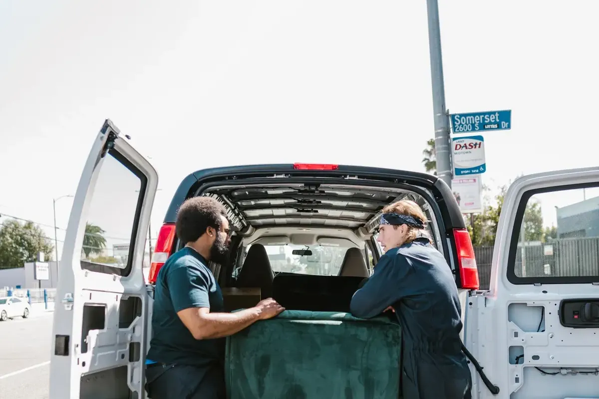 Two people lifting a green sofa into the back of a white van with its rear doors open, parked at the kerb on a sunny street.