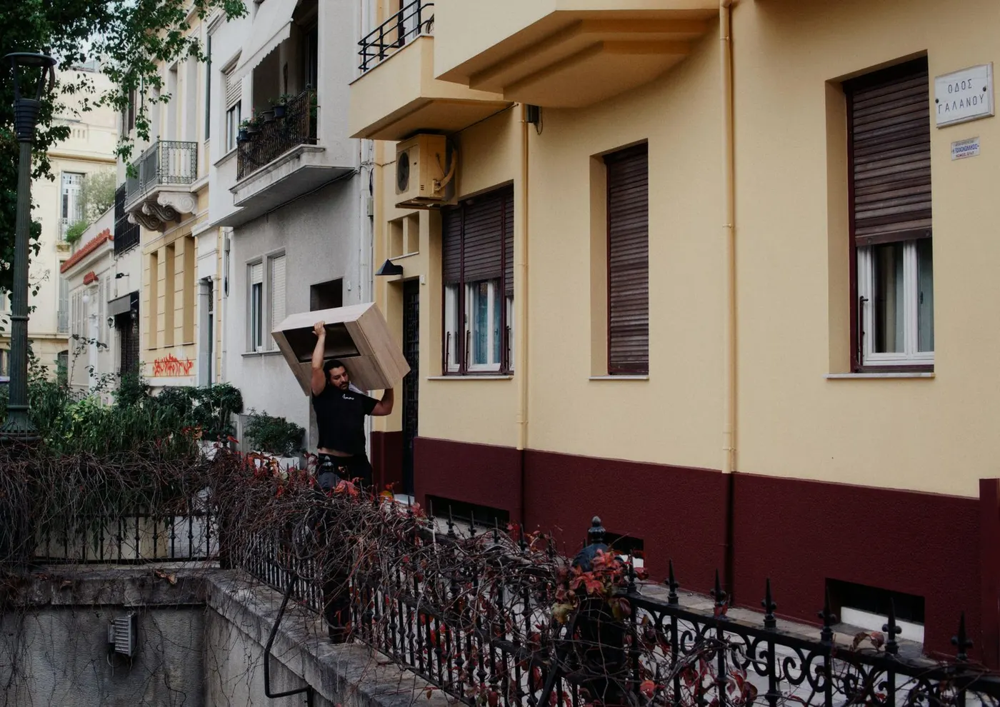 A man carrying a wooden cabinet balanced on his shoulder along a residential street lined with apartment buildings.