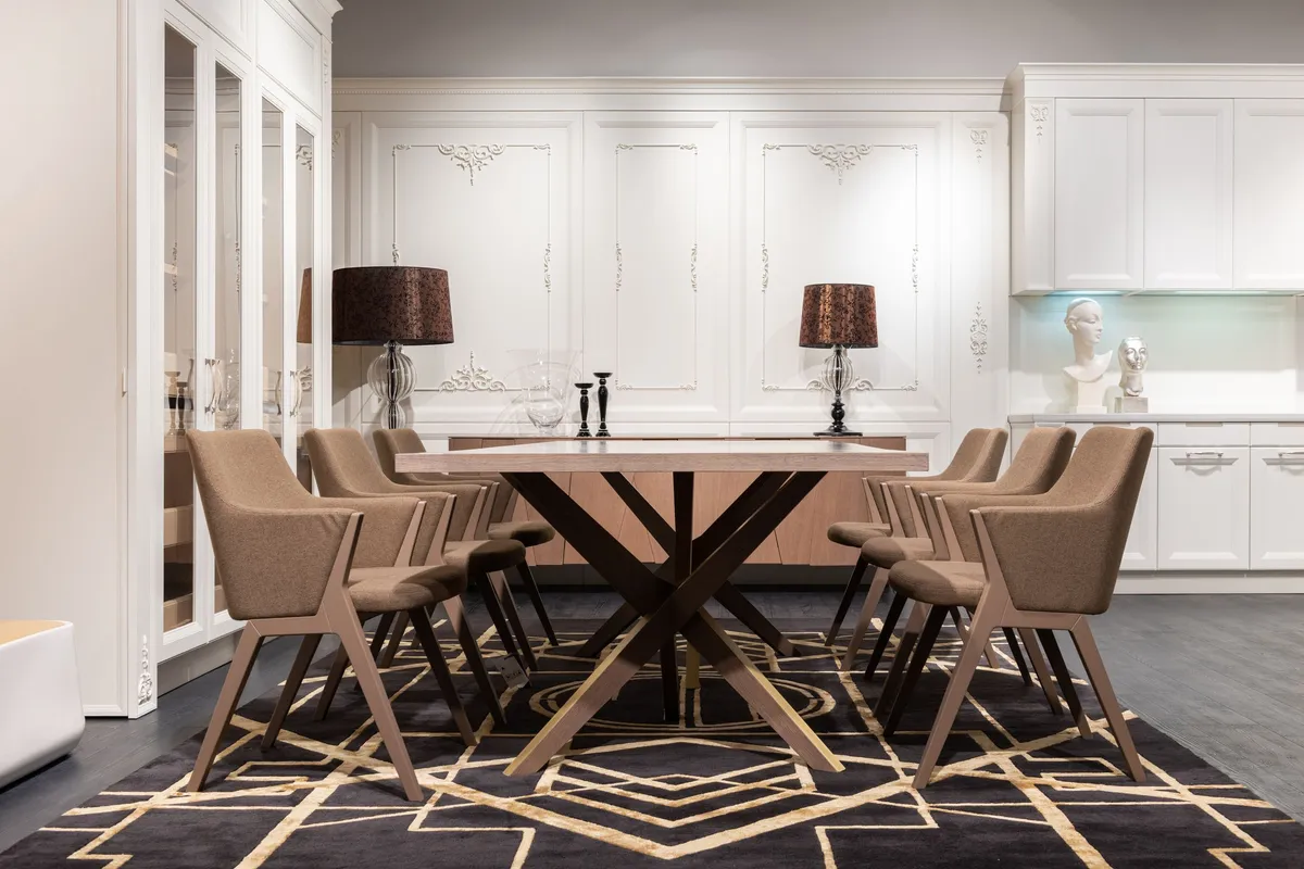 A rectangular wooden dining table on a cross-shaped base surrounded by eight matching fabric-upholstered chairs on a patterned rug.