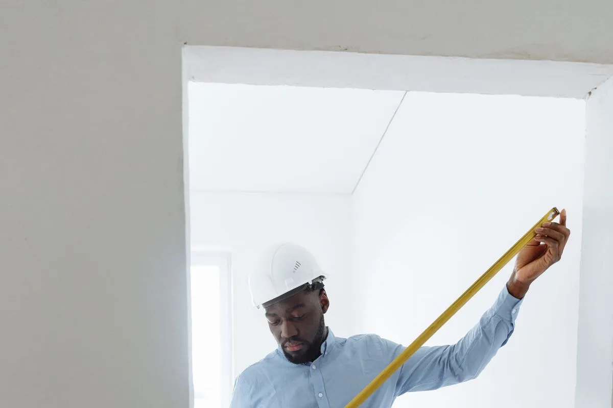 A man in a white hard hat holding a yellow tape measure extended across the width of a doorway opening in a bare, unfurnished room.