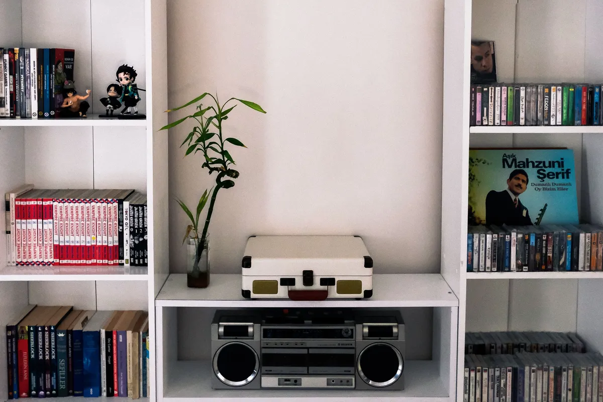 A white media wall unit with open shelving holding a portable record player, a silver stereo system, rows of CDs and DVDs, and books on the side shelves.