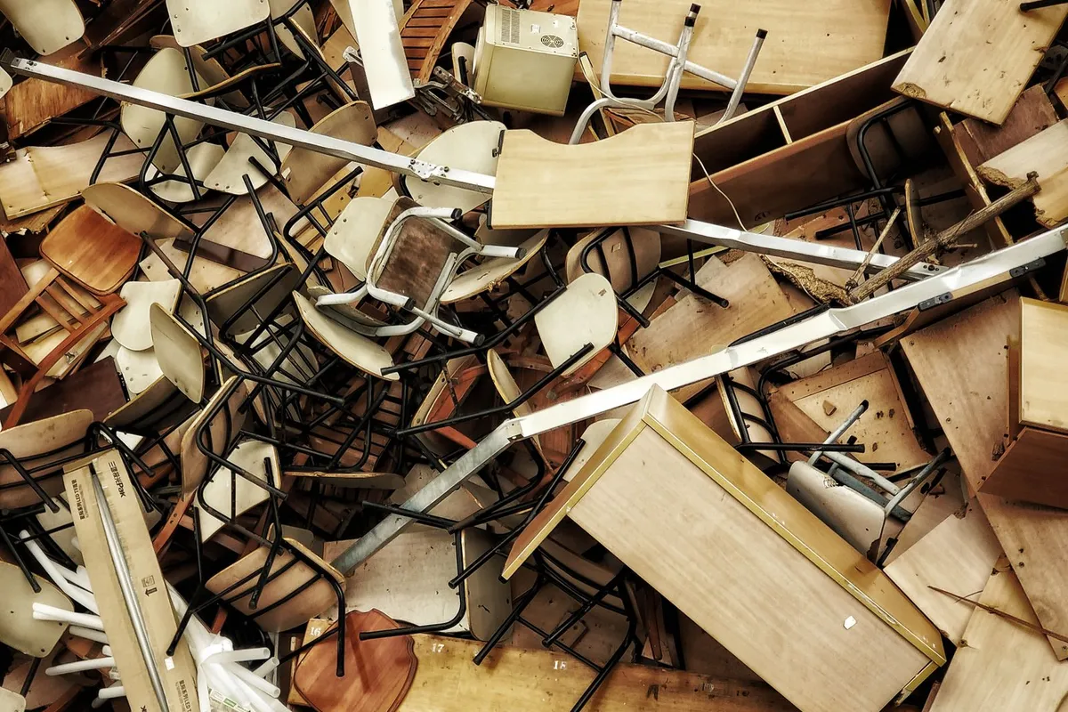 A jumbled overhead pile of broken wooden chairs, desks and drawer units with visible metal legs, fittings and packaging strapping mixed throughout.