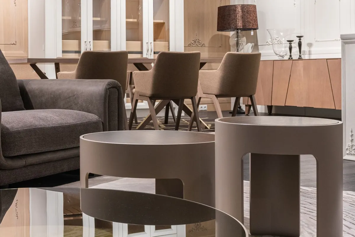 Two round taupe nesting tables of different heights standing one behind the other beside a dark grey sofa, with a low dark glass-topped table in the foreground.
