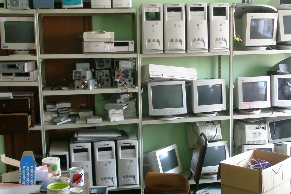 Metal shelving stacked with retired desktop computers, tube monitors, printers and loose power supplies in a storeroom.