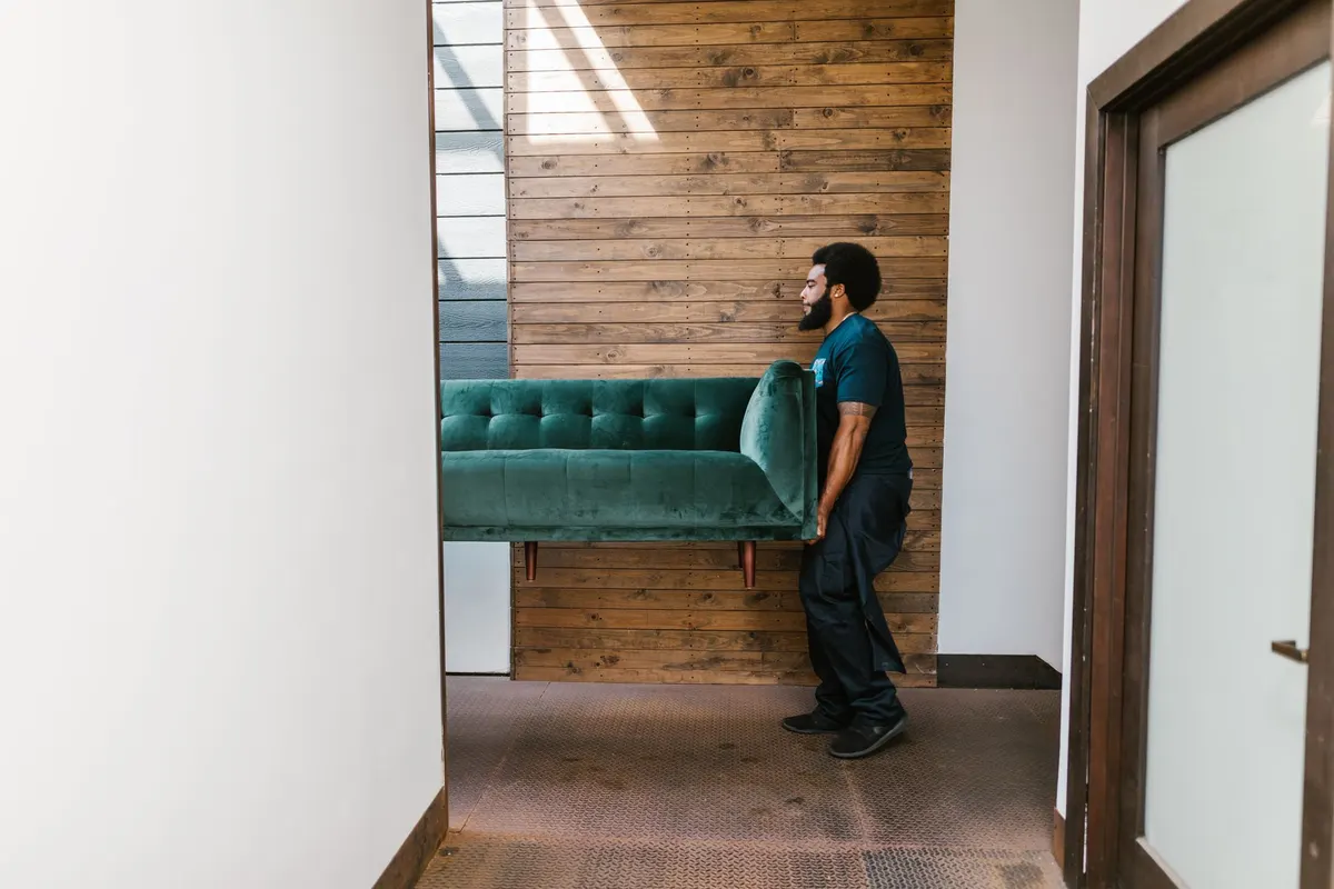A worker in dark work trousers carrying a green velvet sofa on his own along an interior corridor.