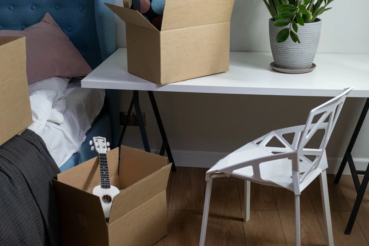 Open cardboard cartons part-filled with belongings on a bedroom floor, beside a white chair, a desk and a made bed.