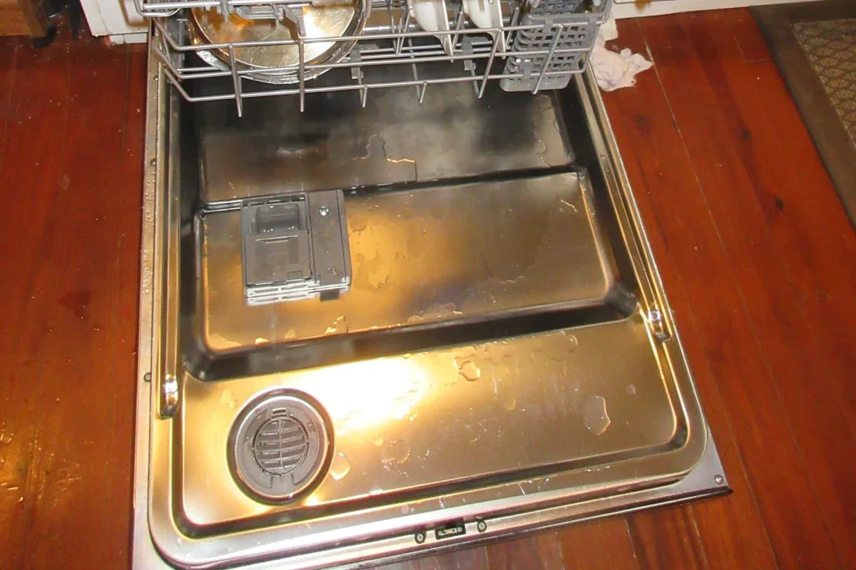 An open dishwasher door showing the lower rack, the stainless steel base of the drum and the circular sump filter set into it.