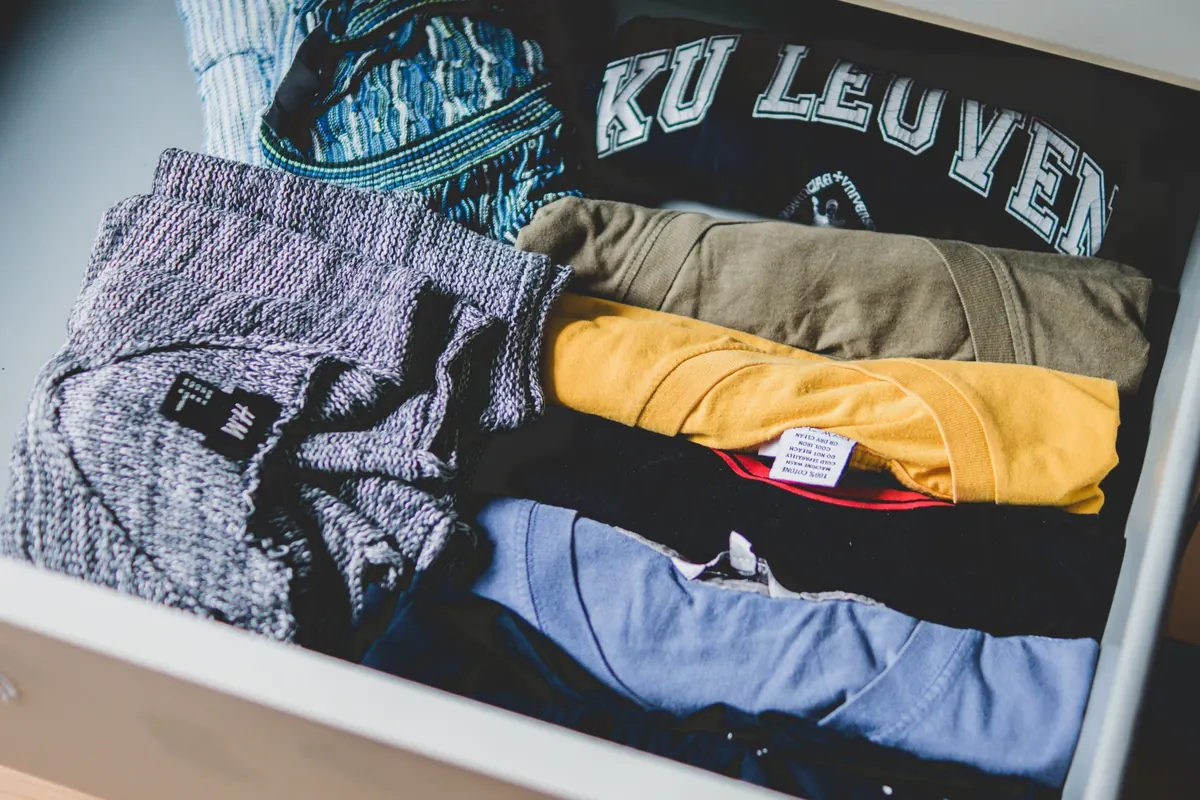 An open drawer of a white chest filled with folded jumpers, T-shirts and jeans, seen from above.