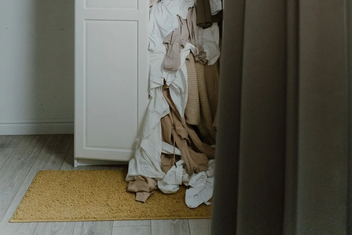 An open white wardrobe with clothes and linen spilling out onto a rug and wooden floor in a bedroom.