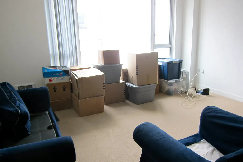 Packed cardboard boxes and plastic storage tubs stacked by the window of a high-rise apartment living room being emptied.