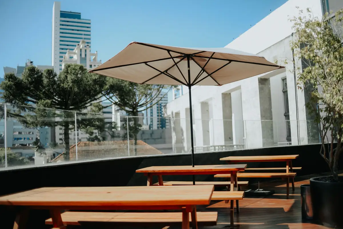 An open cream parasol on a roof terrace, its pole standing in a low black weighted base plate beside wooden benches, with tower blocks behind.