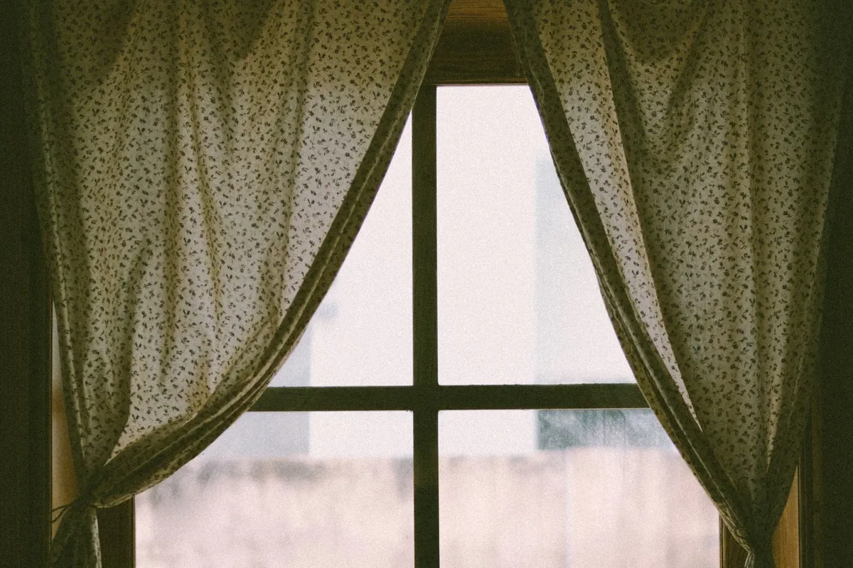 Floral patterned fabric curtains tied back on either side of a paned window, with a small lamp and a drink on the windowsill below.