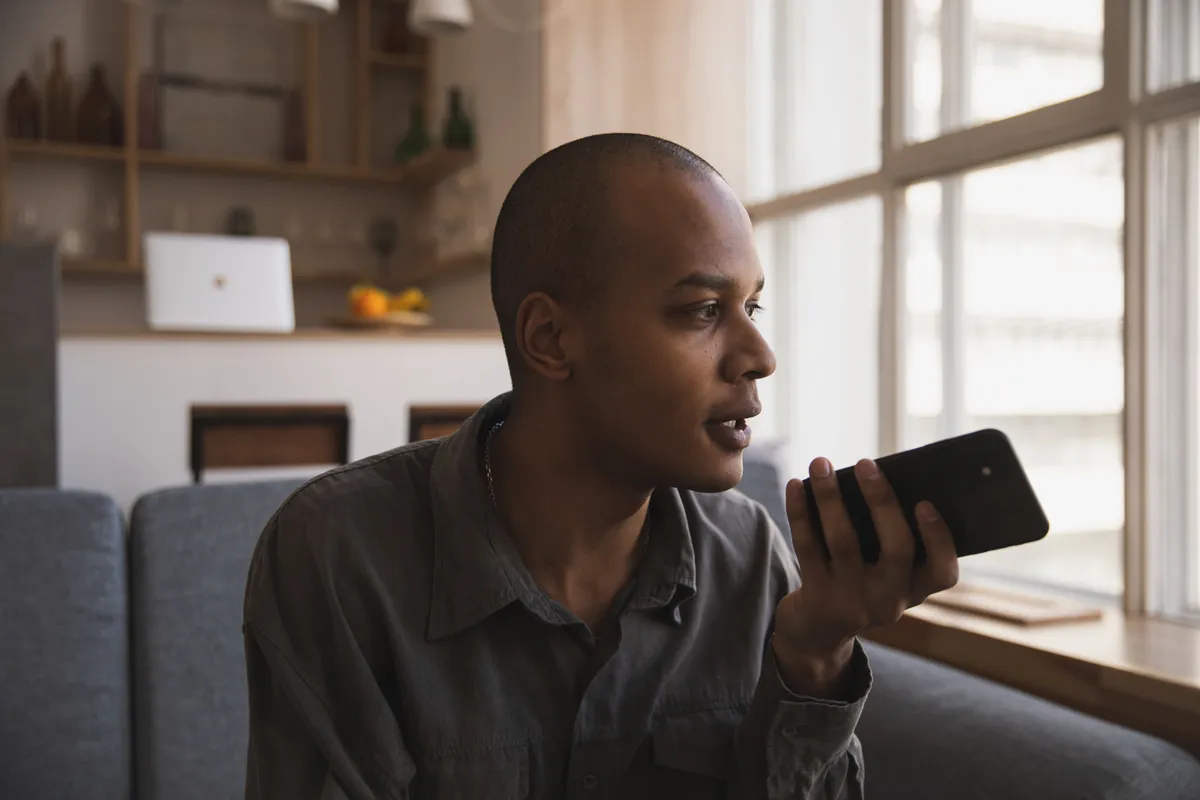 A man sitting on a sofa in a bright living room, speaking into a phone held near his mouth.