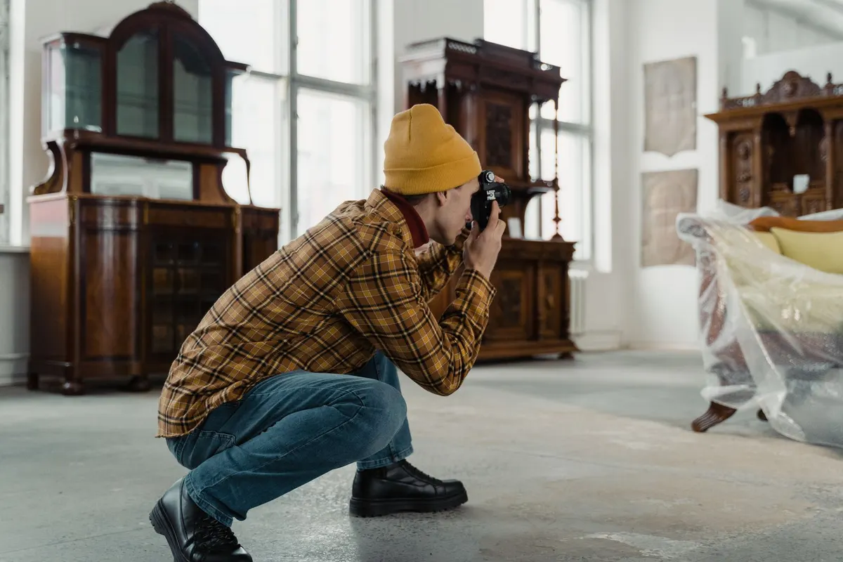 A person crouching with a camera to photograph furniture in a bright, largely empty room, with a plastic-covered sofa in the background.