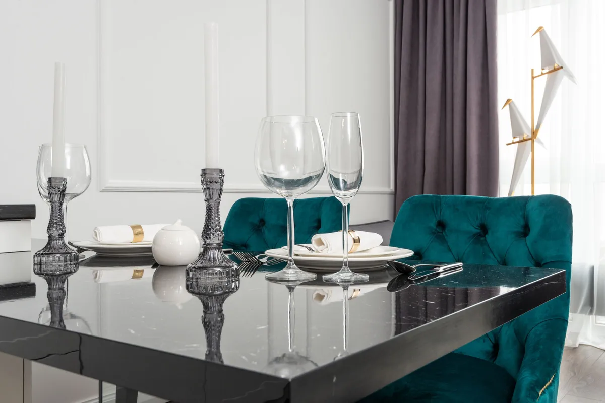 The corner of a polished dark marble dining table with a thick edge, set with plates, glasses and candlesticks beside a green velvet chair.