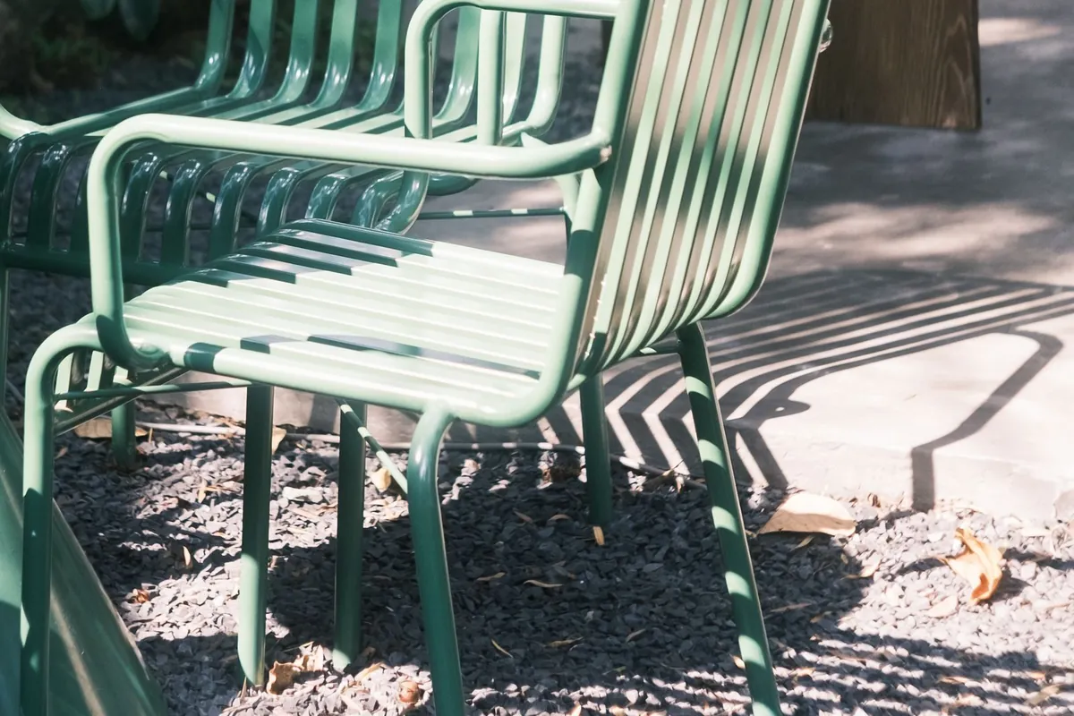 Two green powder-coated metal garden chairs with slatted seats and a matching round table, standing on gravel in dappled sunlight.