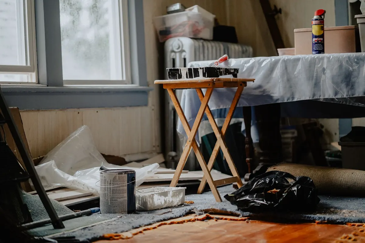 A table covered with a plastic dust sheet beside a folding stand, paint tins and a ladder in a room being prepared for renovation.