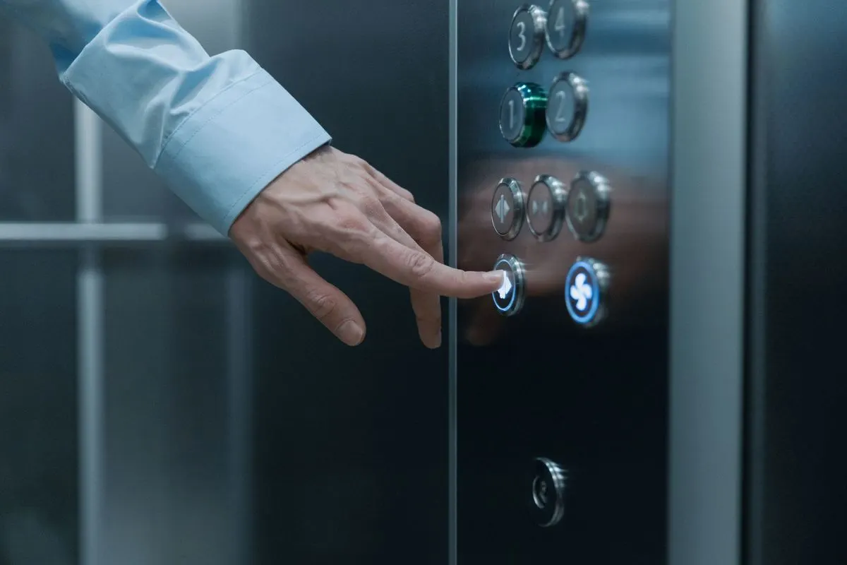 A hand in a light blue shirt cuff pressing an illuminated call button on a stainless steel lift panel inside a building.