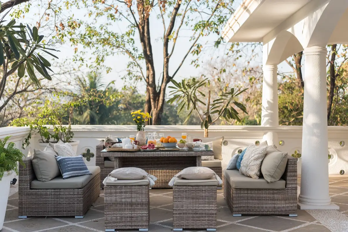 A woven rattan outdoor lounge set with cushioned sofas, two cube stools and a low table on a tiled villa terrace, beside a white balustrade and column.