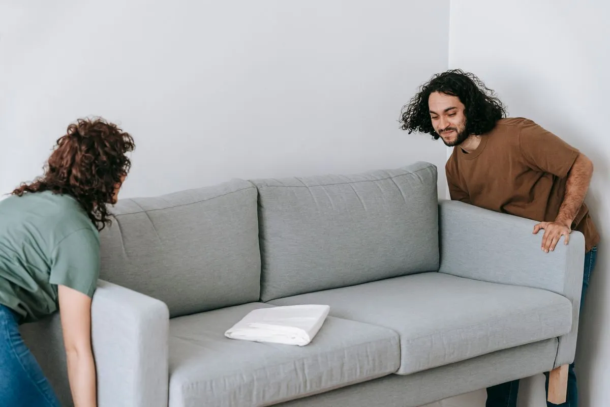 Two people repositioning a grey sofa inside a living room.