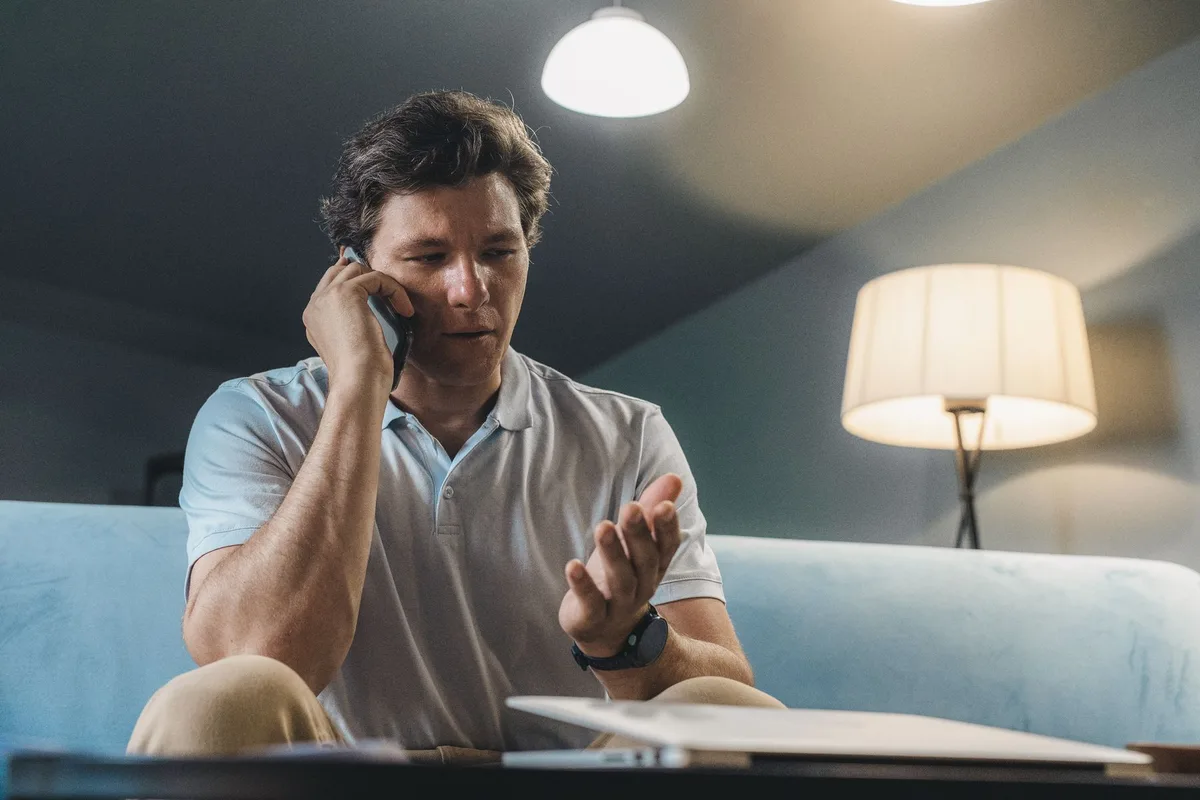 A man sitting on a sofa at home speaking on a mobile phone with papers open on the table in front of him.