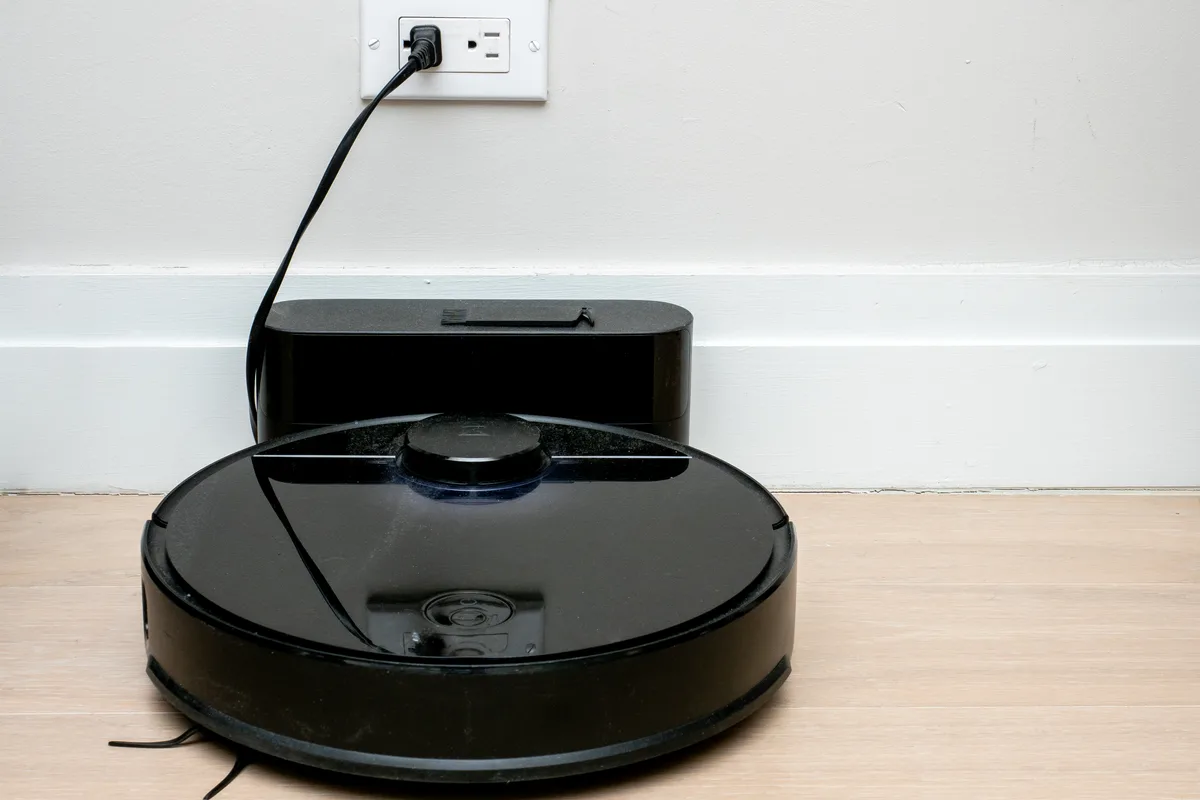 A black robot vacuum cleaner parked on its charging dock against a white skirting board.