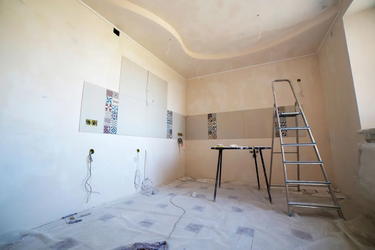 A stripped interior mid-works with dust sheets across the floor, a stepladder, a trestle table, exposed wiring and tiles fixed to a half-finished wall