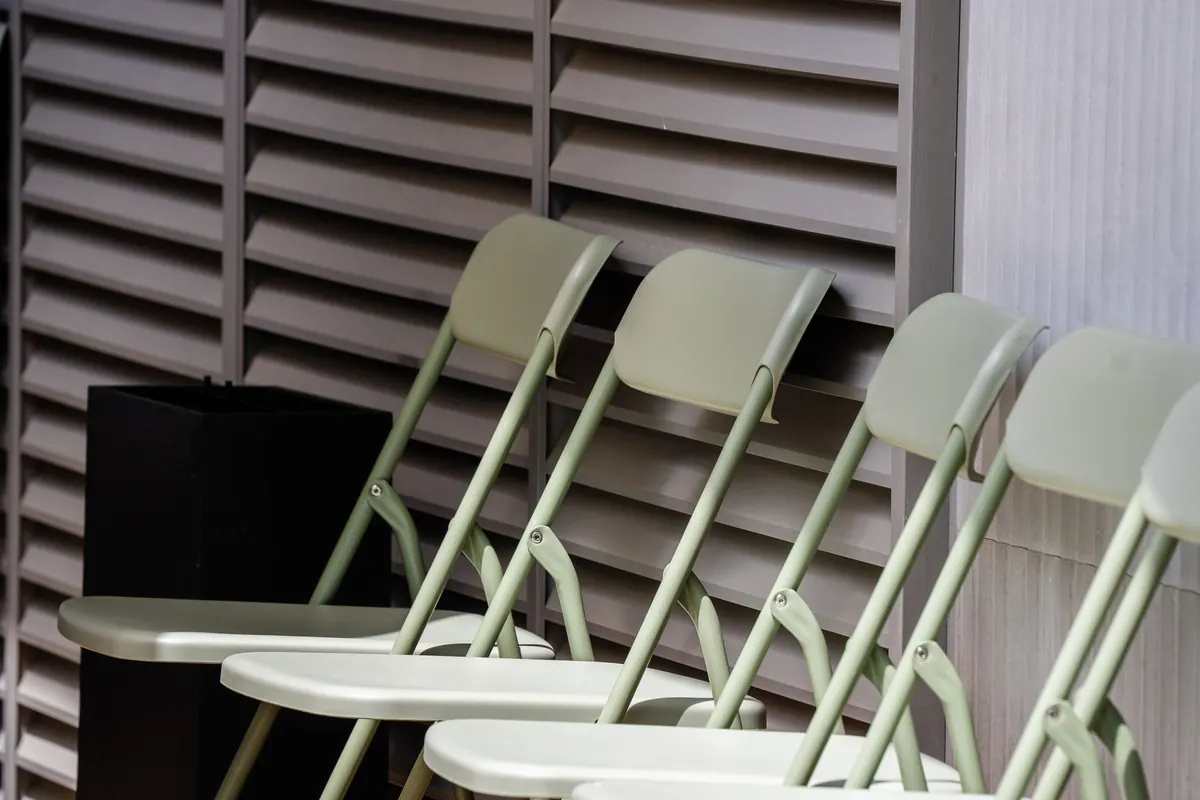 A row of pale green folding chairs standing against a louvred wall, with the hinged tubular frames and pivot rivets clearly visible.