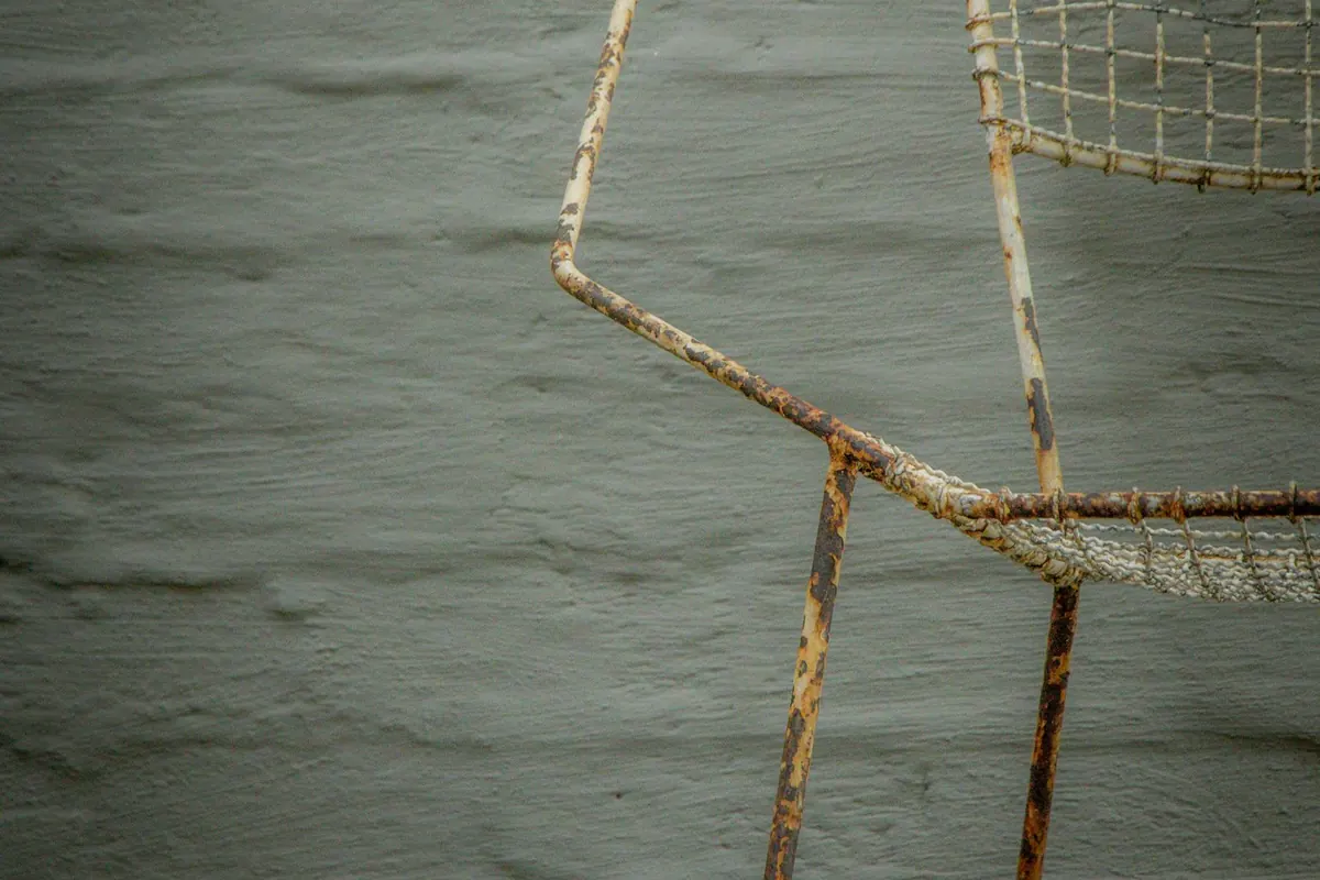 A steel outdoor chair frame against a painted wall, the white paint flaking off the tubing and rust spreading along the arm and down the legs.