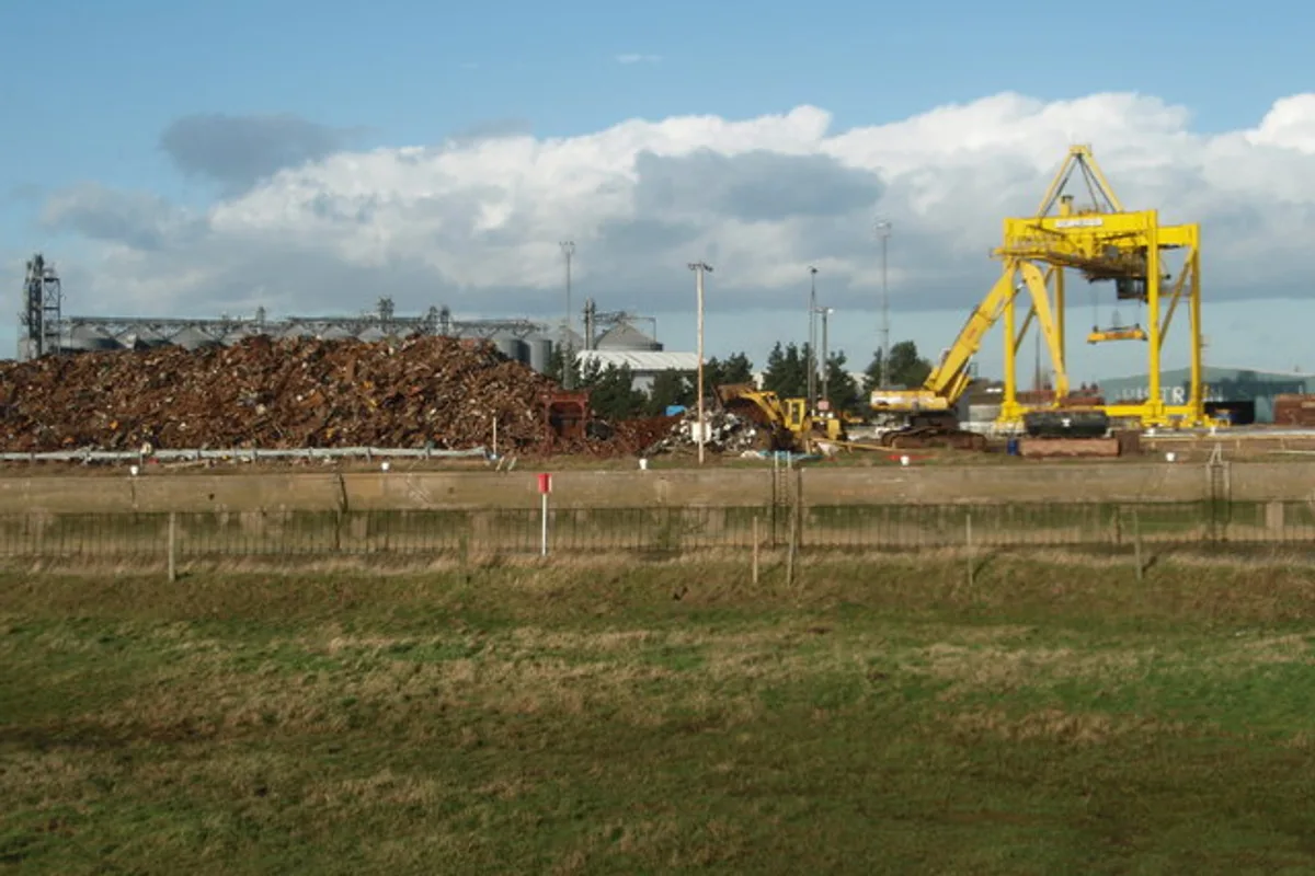 A large open-air scrap metal yard with a crane and machinery beside a tall pile of steel scrap awaiting processing.