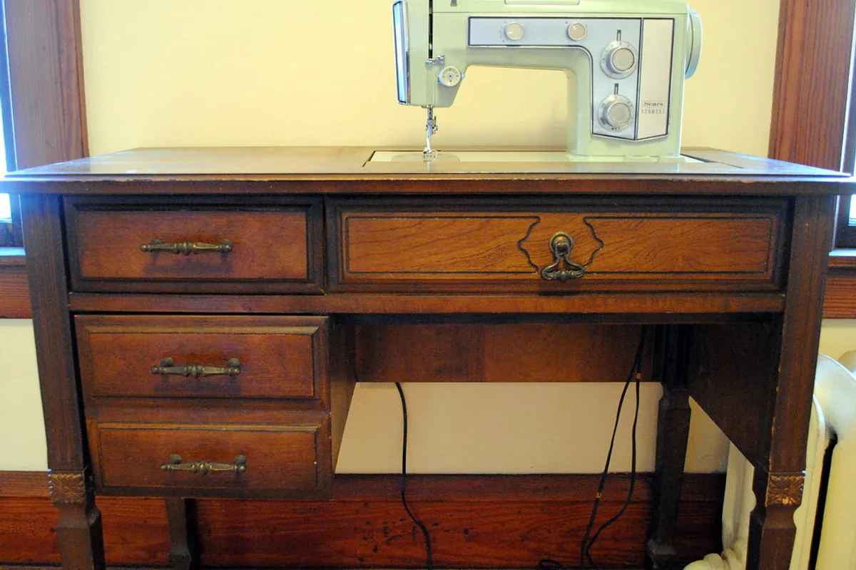 A sewing machine set into the top of a wooden cabinet with brass-handled drawers and a foot pedal cable trailing beneath it.