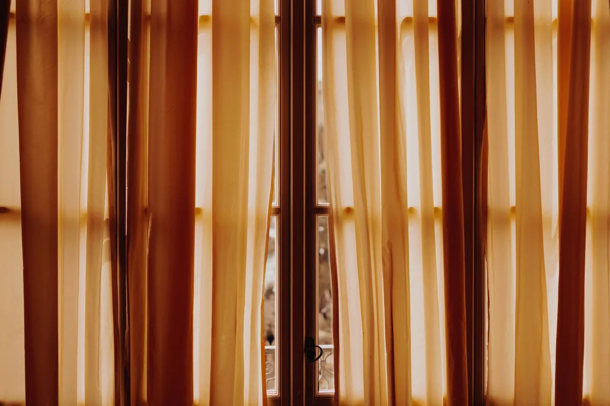 Sheer orange curtain panels hanging in front of a glass door, backlit by strong afternoon sunlight streaming through the fabric.