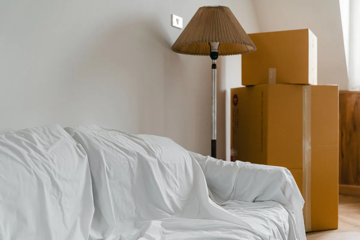 A sofa draped in a white dust sheet beside a floor lamp and a stack of sealed cardboard cartons in a bright, half-empty room.