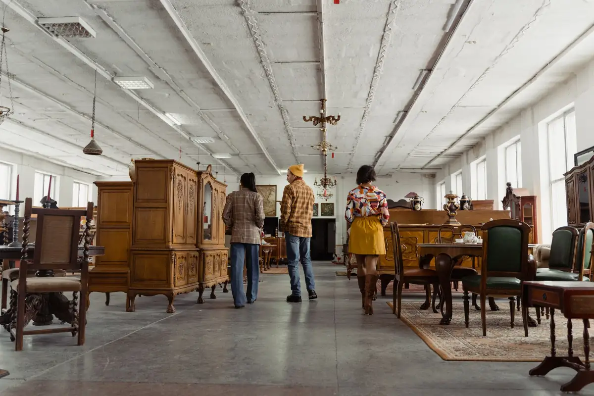 Three shoppers walking between wooden cabinets, dining tables and upholstered chairs in a bright second-hand furniture showroom.