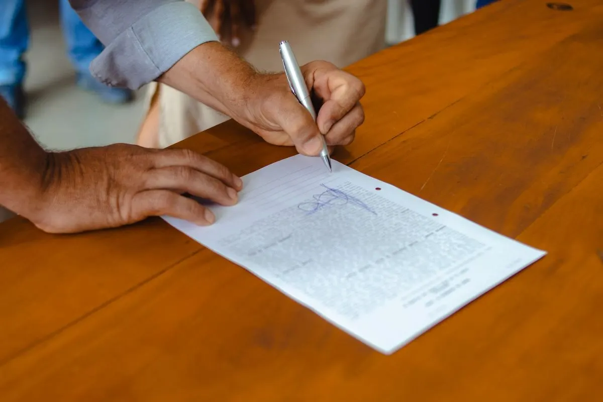 Two people at a wooden counter, one steadying a printed one-page form while the other signs it with a pen.