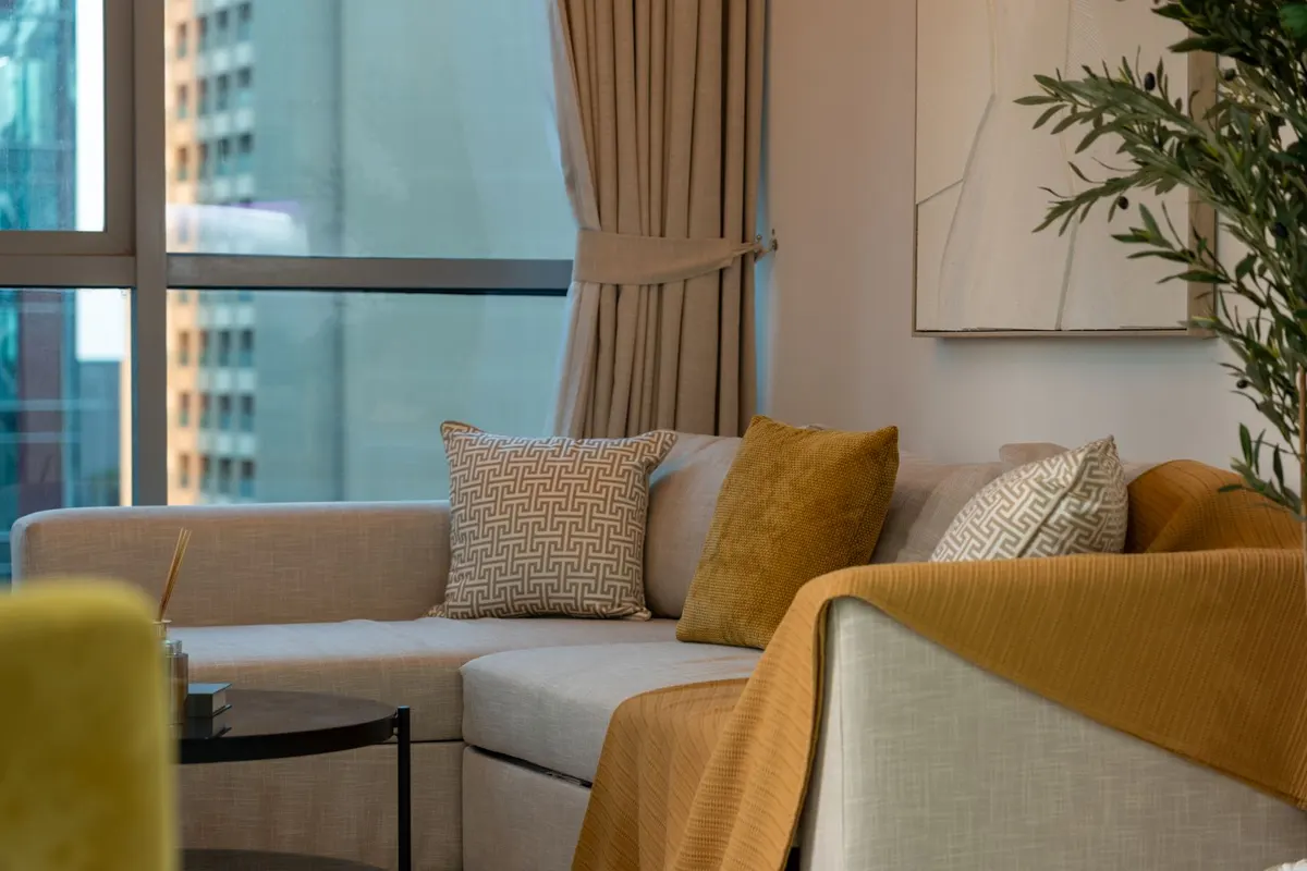 A beige fabric sofa with patterned cushions and a mustard throw in a living room, beside a floor-to-ceiling window looking out at a residential tower.