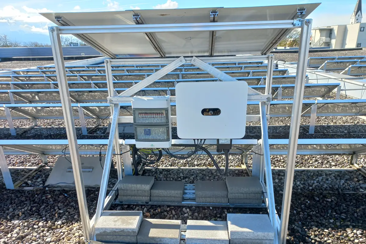 An inverter box and a DC isolator mounted on the aluminium frame beneath a rooftop solar panel, with concrete ballast blocks holding the frame down.