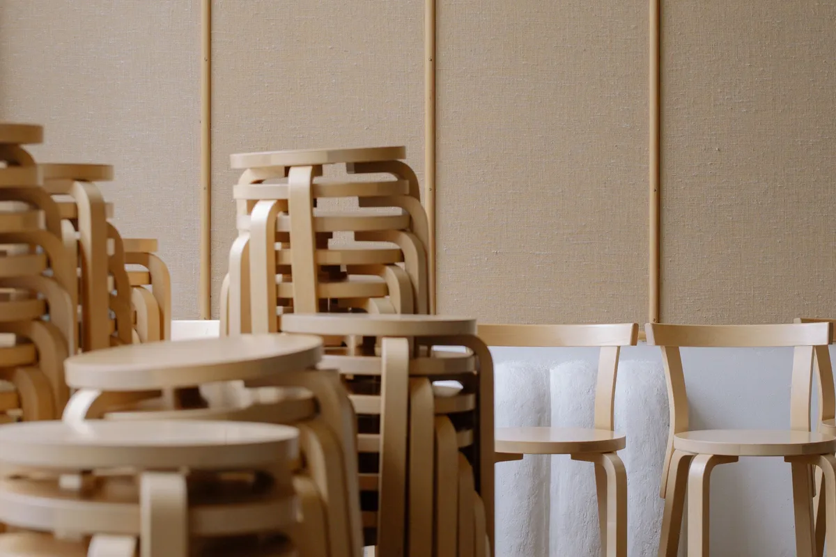Stacks of light birch chairs piled four and five high beside two single chairs standing against a fabric-panelled wall.