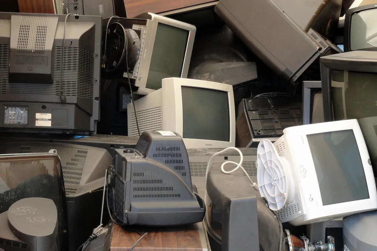 A dense stack of dozens of old televisions and computer monitors of different sizes piled together, with power leads trailing between them.