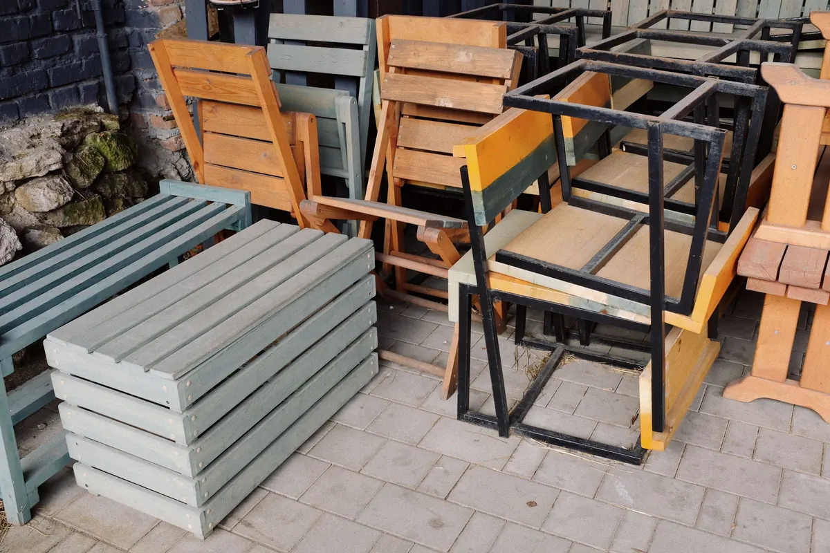 Wooden benches, folding chairs and stacked timber frames set aside together on a paved area beside a wall.