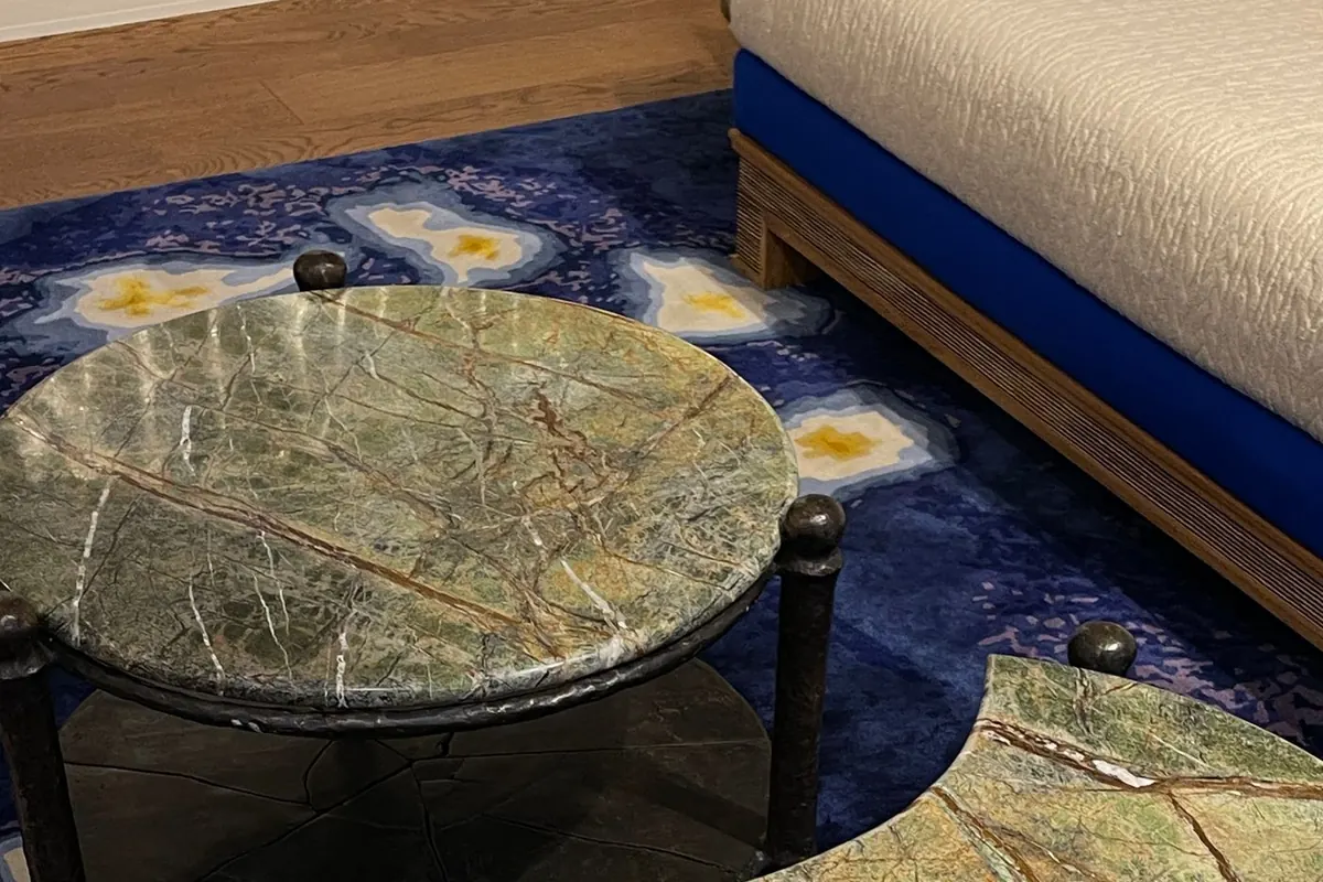 A round green marble table top with dark veining resting on a slim dark metal frame, with a second matching stone-topped table beside it on a blue patterned rug.