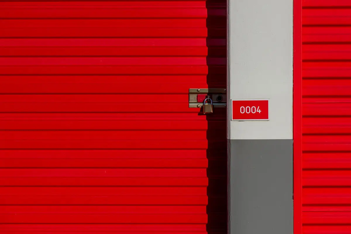 A red roller shutter door of a self-storage unit secured with a brass padlock, next to a numbered unit sign reading 0004.
