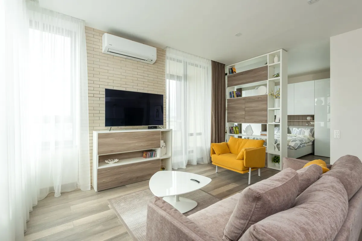 A studio apartment interior with a sofa and television in the foreground and a bed visible through an open partition behind a yellow armchair.
