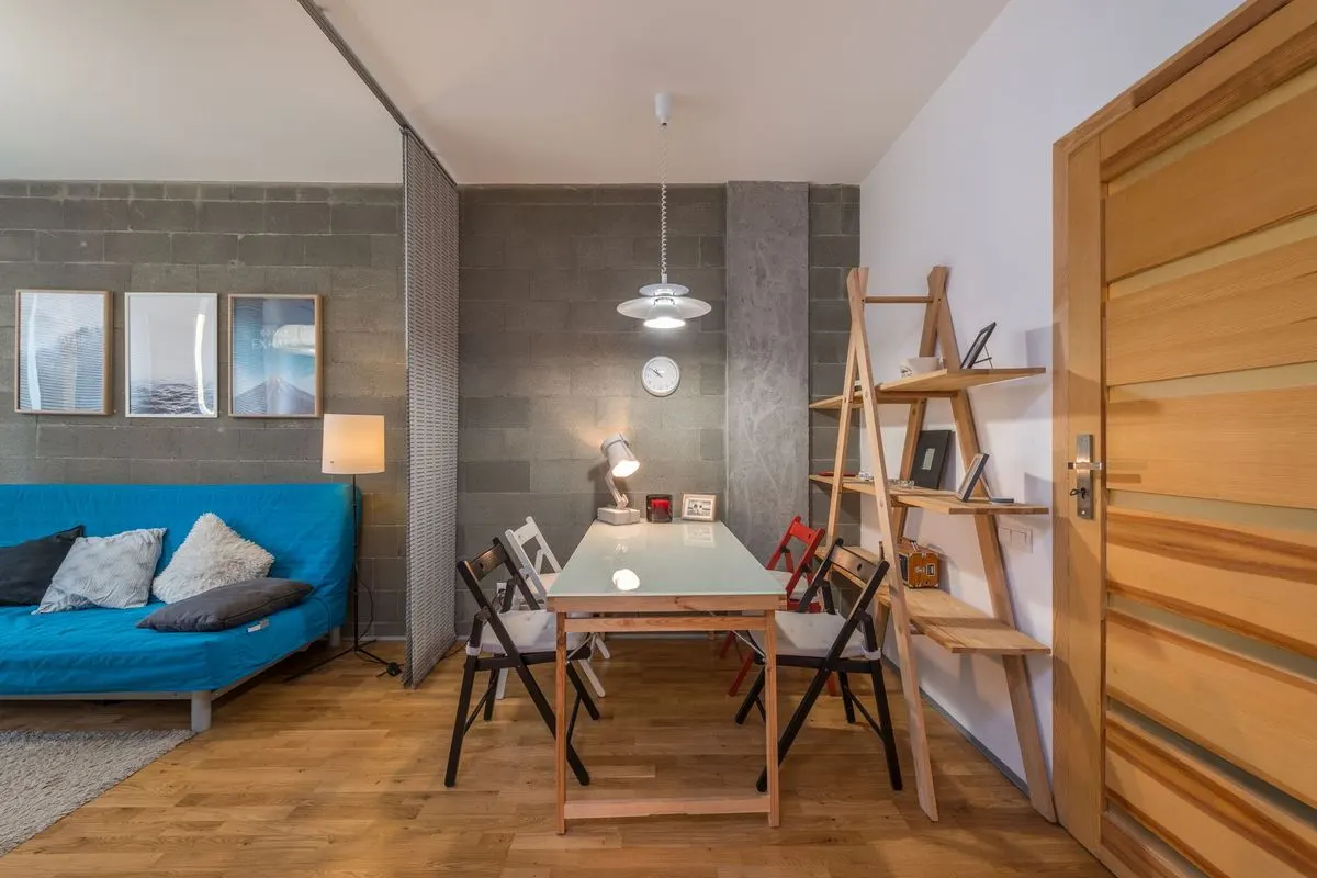 A studio living space holding a blue sofa bed, a glass-topped table with folding chairs, a leaning timber shelf unit and a rug.