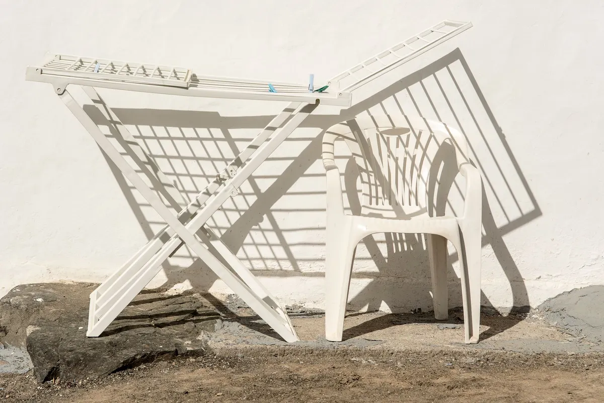 A white folding plastic clothes-drying rack and a white plastic chair standing against a sunlit white wall, casting hard shadows.