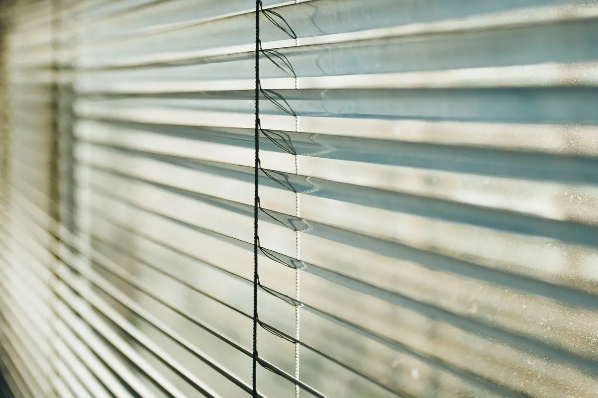 Sunlight streaming through the angled metal slats of a venetian blind, casting striped shadows and highlighting the lift cords running through it.