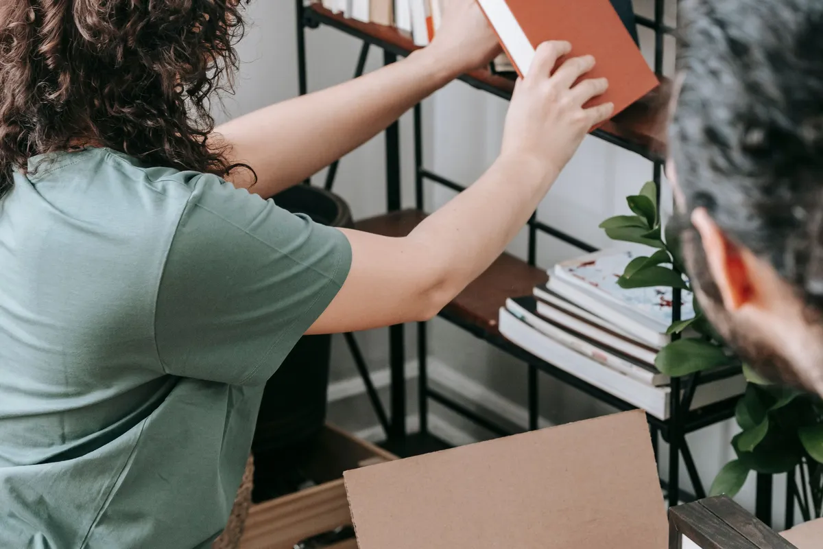 A person lifting a red hardback book from an open metal and timber shelving unit, with an open cardboard box in the foreground.