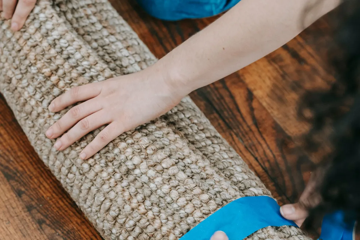 Two people rolling a jute rug tightly on a wooden floor and securing it with a strip of tape.